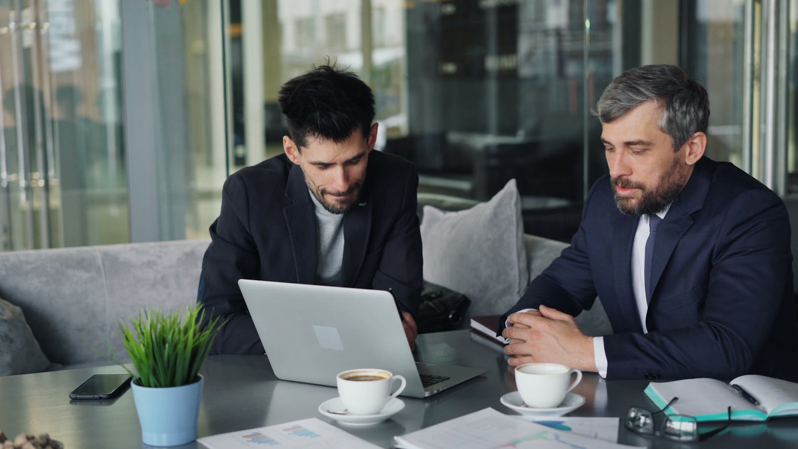 Two businessmen in a modern cafe collaborating over laptops and coffee.