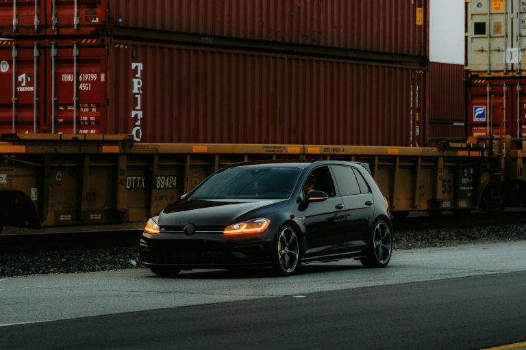 A black car drives along a road, passing large stacked cargo containers.